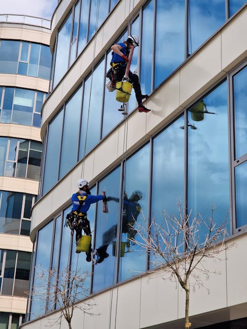 Two professional window cleaners from Hammersmith Cleaners are suspended on harnesses high above the ground, meticulously cleaning large glass windows of a modern multi-storey building in Hammersmith.