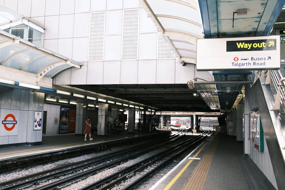 A modern glass office building with reflective surfaces showing neighboring structures, standing behind the historic brick Hammersmith station façade with a clock and station sign, set against a pale